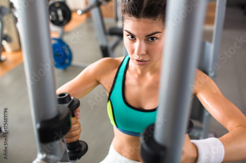 Confident athletic woman looking at camera while having intensive training on butterfly machine at modern gym, waist-up portrait