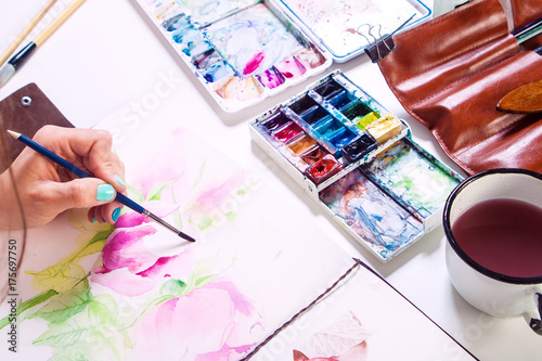 Close-up of a woman artist paints watercolor drawing of a pink flower of a peony and a dog rose in the album for drawing on a white table, beside lies a watercolor palette and a cover with tassels