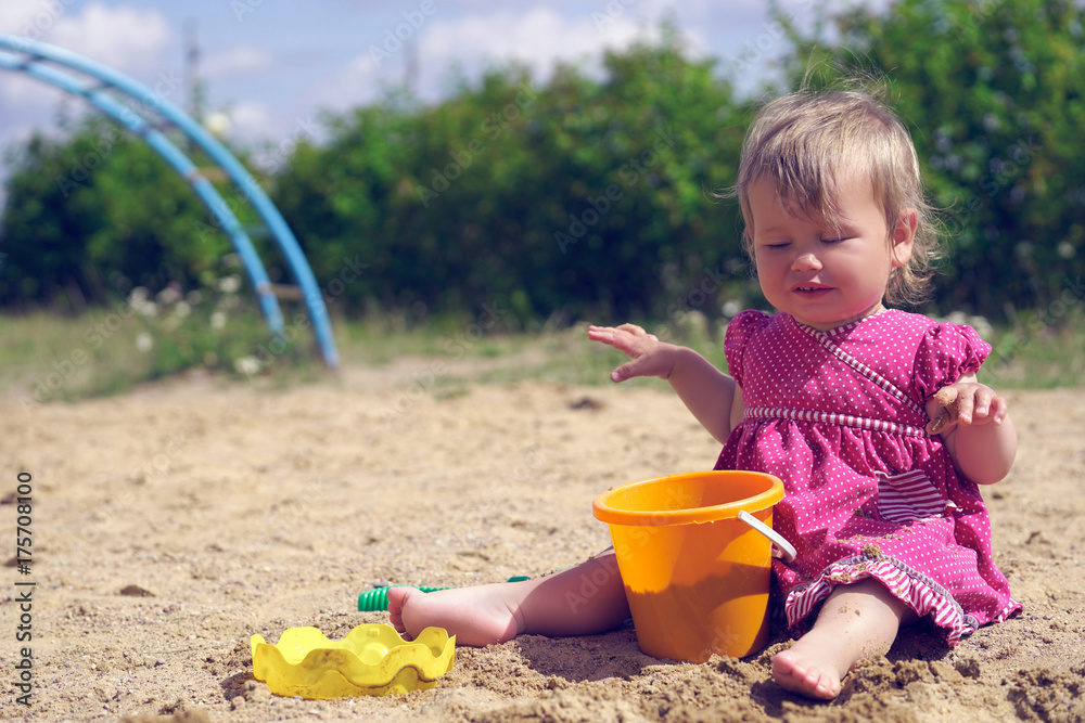 A little girl squints against bright sunlight. Adorable toddler girl in dress play with sand on sandbox. A little girl squints against bright sunlight. Adorable toddler girl in dress play with sand on sandbox.