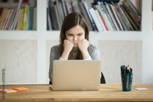 Grumpy not amused office worker looking at laptop screen in office. Young business lady bored with mundane workflow, tired from working too much. Casual businesswoman not happy with boring work task.