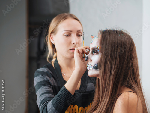 Make-up artist make the girl halloween make up in studio.Halloween face art.Woman applies on professional greasepaint on the face of girl.War-paint with blood, scars and wounds. photo