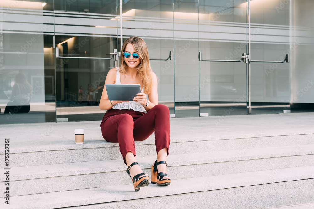 Beautiful young woman sitting in the stairs on the tablet wearing sunglasses