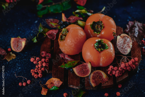 Ripe persimmons on wooden plate in a dark autumn still life with fruits and berries. Harvest concept. Dark food photography with copy space. photo