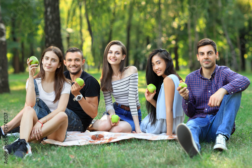 Portrait of cheerful friends holding green apples sitting on grass