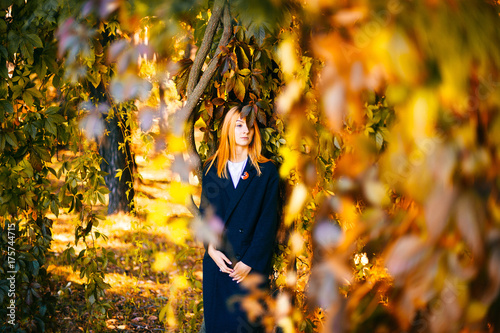 Lonely sad young cute beautiful girl with red hair and reflexive face standing beyond tree in botanical garden. Yellow colors in october. Countryside wild terrain. Vegetation, bushes, leaves in autumn photo