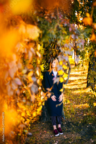 Lonely sad young cute beautiful girl with red hair and reflexive face standing beyond tree in botanical garden. Yellow colors in october. Countryside wild terrain. Vegetation  bushes  leaves in autumn