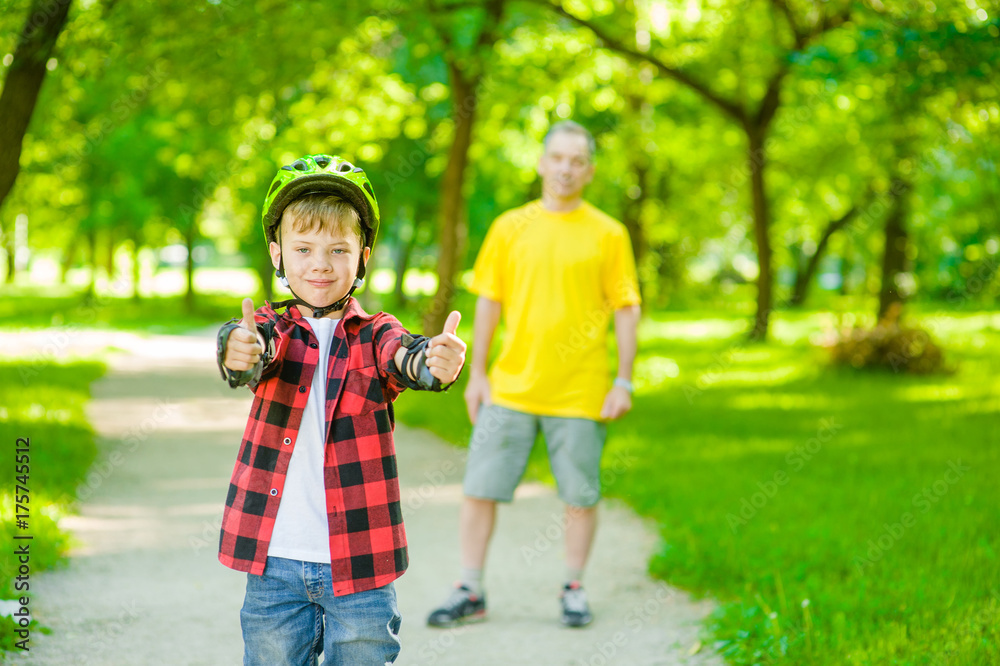 Boy in a protective helmet and protective pads for roller skating showing thumbs up on the background of his father Boy in a protective helmet and protective pads for roller skating showing thumbs up on the background of his father