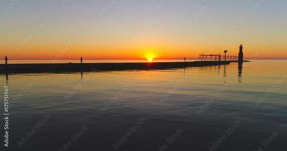 Fishermen, pier, and Lighthouse silhouetted against fiery Lake Michigan Sunrise over tranquil waters, moving low aerial view. Fishermen, pier, and Lighthouse silhouetted against fiery Lake Michigan Sunrise over tranquil waters, moving low aerial view.