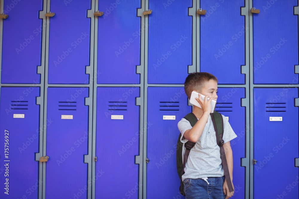 Fotografie Elementary boy talking on mobile phone against lockers