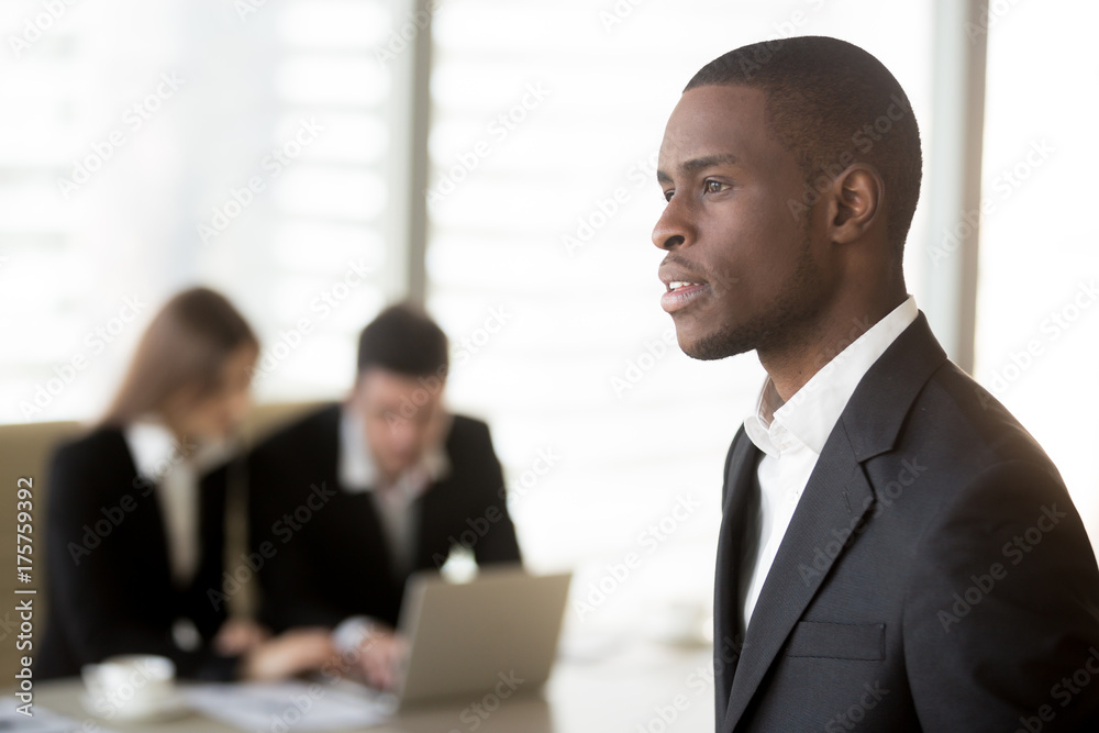 Φωτογραφία Side view portrait of handsome black businessman looking in distance, caucasian businesspersons working on laptop at office desk on background
