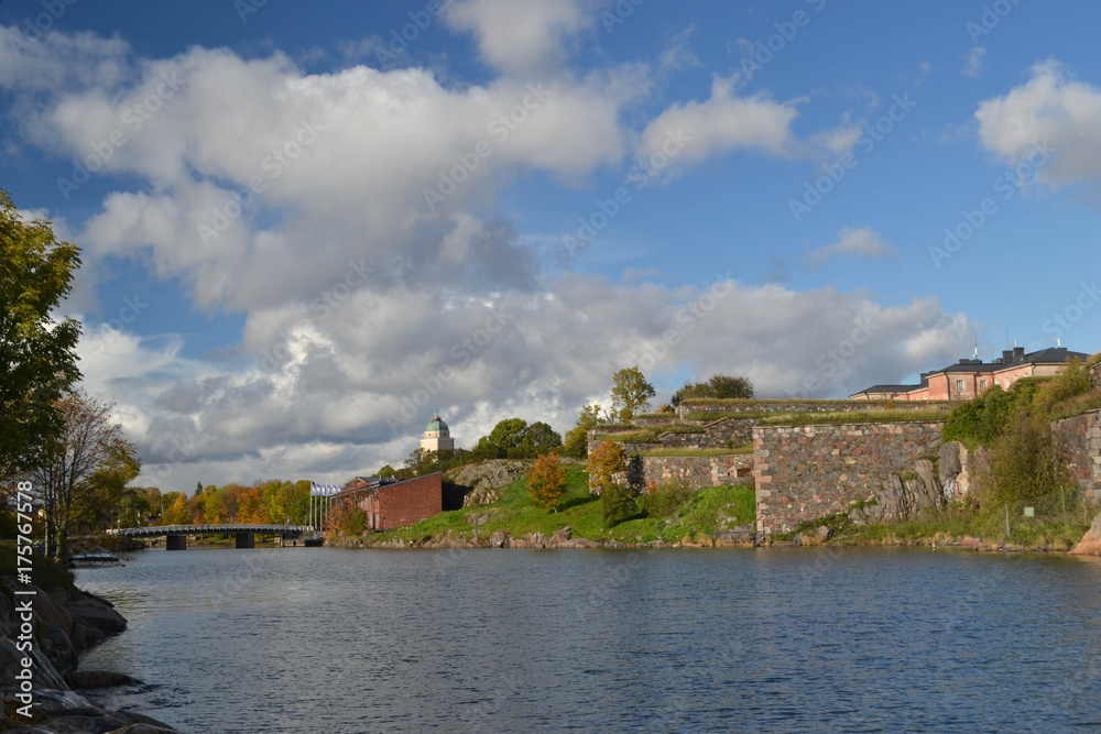 Fototapeta premium Stone Wall by Water with Blue Sky, Bridge, Fluffy Clouds and Fall Color in Background