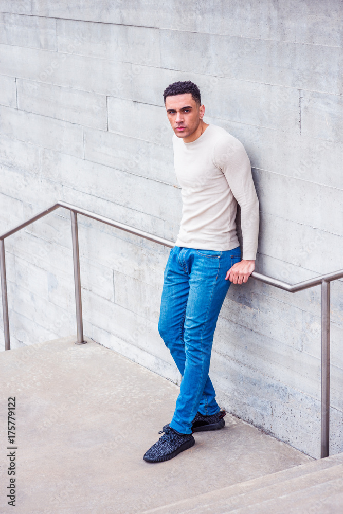 Young American Man wearing light gray collarless, knit pullover sweater, blue  jeans, black sneakers, standing by wall in New York in spring day, looking  up, thinking. Color filtered with purple tint. Stock