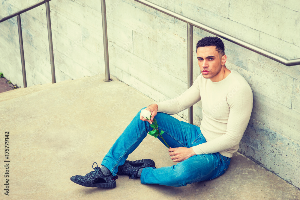 Young American Man wearing light gray knit sweater, blue jeans, black sneakers, hands holding white rose, sitting on ground by wall in corner of street in New York, looking down, thinking, missing you