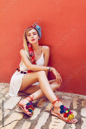 Beautiful woman sitting on a stone street leaning against a red wall. In her beautiful long straight blonde hair is styled colorful scarf. Wearing nice white dress and colorful sandals.