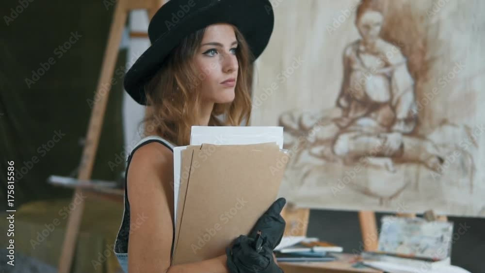 Portrait of lovely female artist holding her sketches and sitting before the easel, slow indoor motion