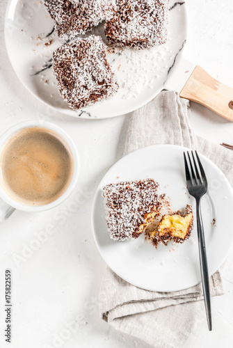 Australian food. Traditional dessert Lamington - pieces of biscuit in dark chocolate, sprinkled with coconut powder chips. On a marble plate, white table. With coffee mug. Copy space top view