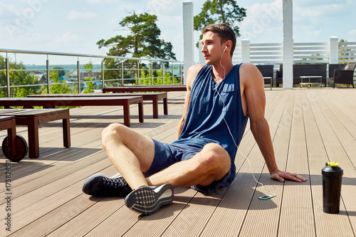 Taking short break from intensive workout: young sporty man enjoying picturesque view while listening to music in headphones and sitting on wooden floor of spacious terrace