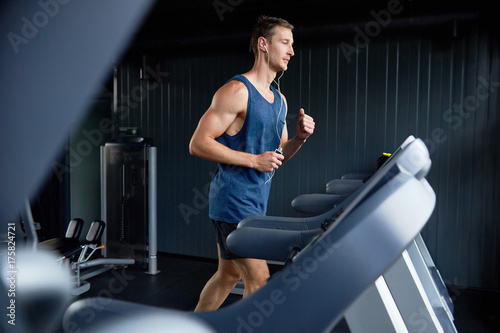 Full length portrait of muscular sportsman listening to music in headphones and using treadmill while wrapped up in training at modern gym, profile view photo