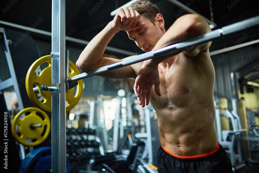 Waist-up portrait of muscular young man with closed eyes leaning on barbell while taking break from intensive workout at spacious gym Waist-up portrait of muscular young man with closed eyes leaning on barbell while taking break from intensive workout at spacious gym