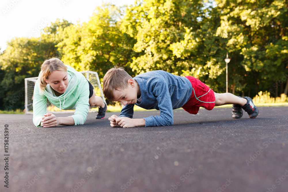 Fototapeta premium Fitness mother with her 9 years old son. Sports mom with kid doing morning work-out at park. Mum and child do the exercises together, healthy family lifestyle concept