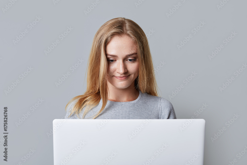 Concentrated female model reads attentively news online, being focused in screen of laptop computer, isolated over grey background. Blonde beautiful young woman surf social networks uses wi fi Concentrated female model reads attentively news online, being focused in screen of laptop computer, isolated over grey background. Blonde beautiful young woman surf social networks uses wi fi