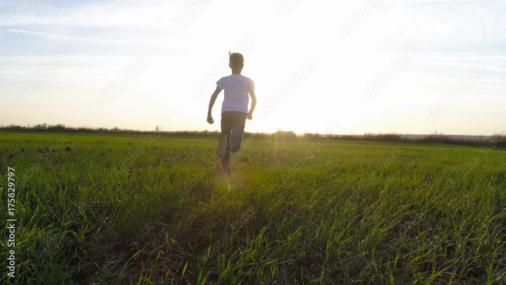 A happy boy in white clothes runs across the green field towards the sun A happy boy in white clothes runs across the green field towards the sun