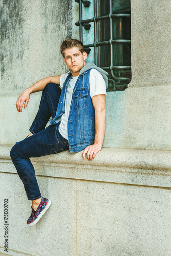 Young American Man with little beard, wearing blue Denim hoody sleeveless vest Jacket, white T shirt, jeans, fashionable shoes, sitting by vintage wall with window on street in New York, relaxing. .