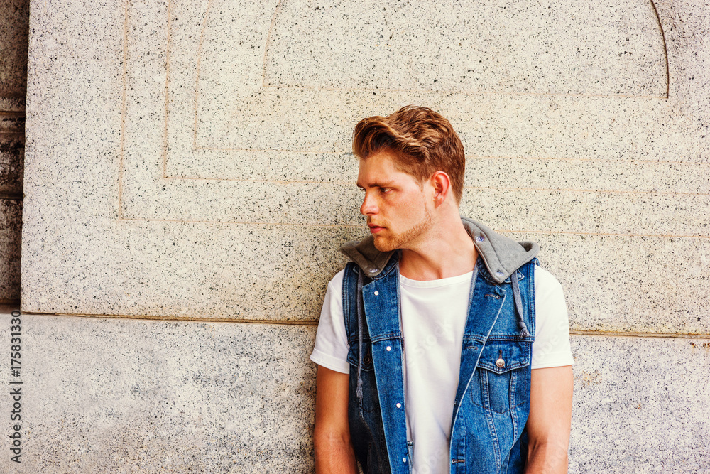 Unhappy Young American College Student with little beard in New York,  wearing blue Denim hoody sleeveless vest jacket, white T shirt, standing  against vintage wall on campus, frowned, sad, thinking. . Stock