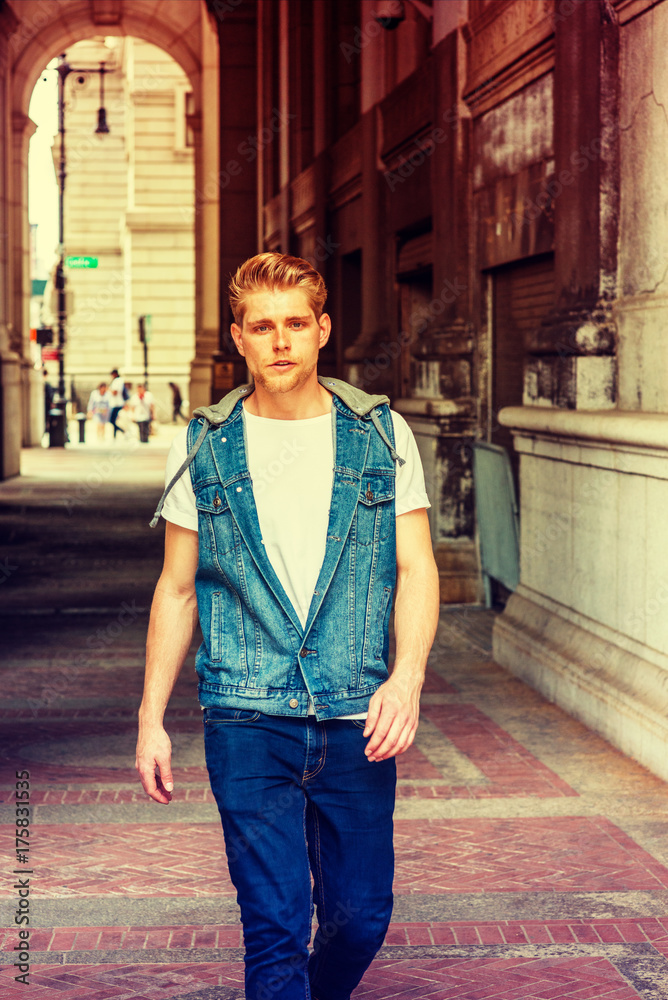 Young American Man with little beard traveling in New York, wearing blue Denim hoody sleeveless vest jacket, white T shirt, jeans, walking through small, narrow vintage street. Color filtered effect.