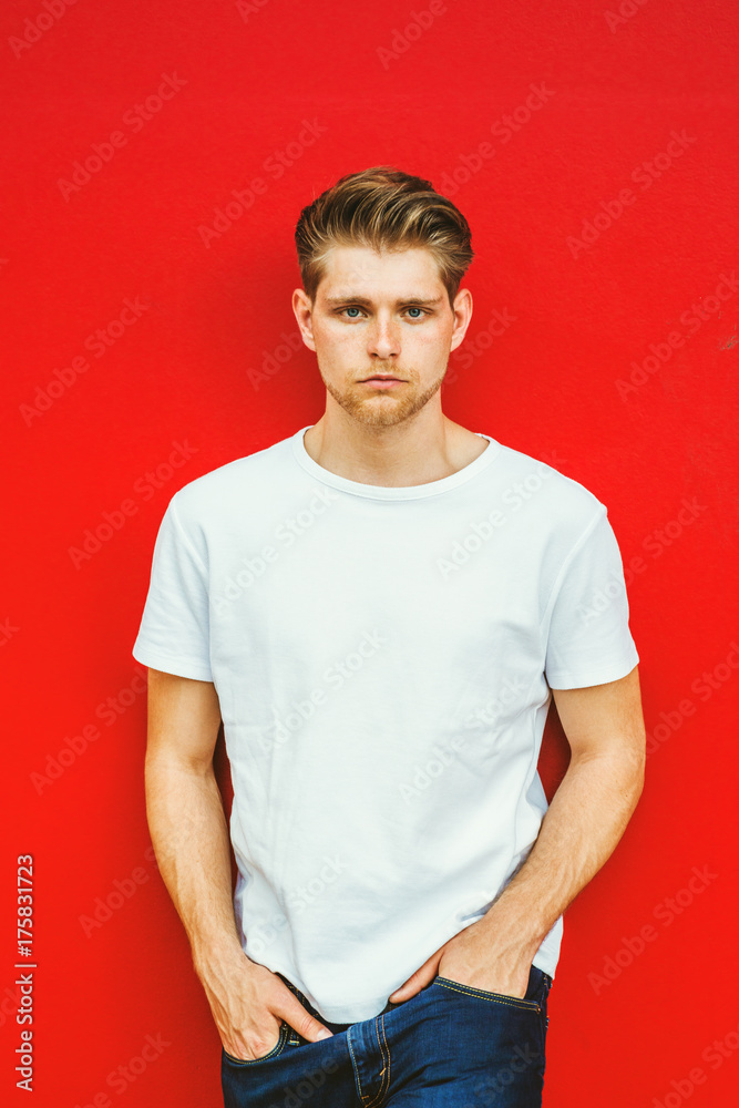 Portrait of Young Handsome Man with little beard in New York, wearing white T shirt, blue jeans, hands in pocket, standing against red background, seriously looking at you.. Portrait of Young Handsome Man with little beard in New York, wearing white T shirt, blue jeans, hands in pocket, standing against red background, seriously looking at you..