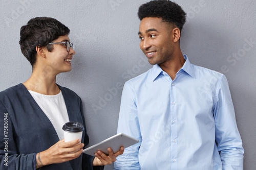 Sophisticated mixed race female and male colleagues have conversation together, discuss future meeting with partners. Elegant brunette business lady holds tablet and coffee meets young boss at office