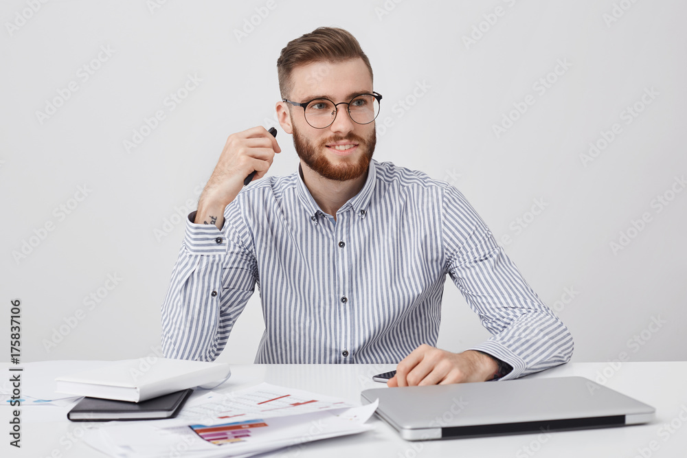 Fototapeta premium Portrait of male office worker, has specific appearance, dressed formally, sits at white desk, uses laptop, smart phone and documents for work, looks aside as notices his best companion or colleague
