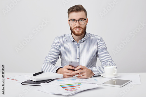 Isolated stdio shot of pleasant looking stylish male manager with stubble, holds smart phone as messages online, uses high speed internet connection, drinks coffee, rests after studying documents photo