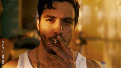 Smoking Brutal Gang Member with Gold Chain Looks into Camera with Defiance. Underground Drug Laboratory is in Background.