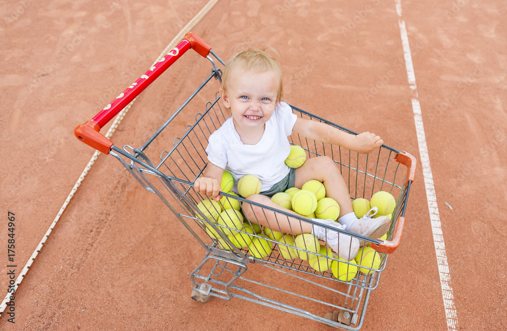 The little girl sits in a basket for tennis balls. View from above