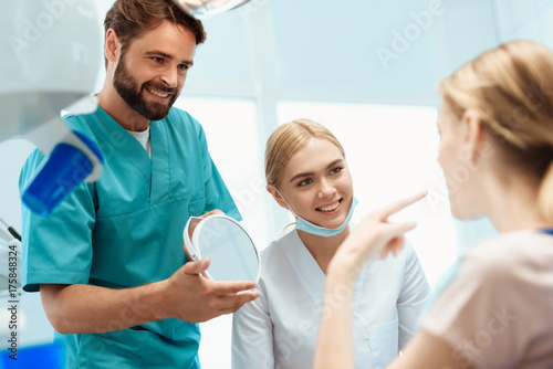 A woman is sitting in a dental chair. Before her stands a doctor and holds a mirror in her hands. Near the nurse