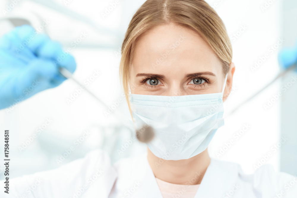 A dentist in a white medical mask is holding a dental mirror and a probe