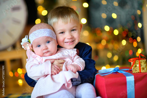 brother hugs his younger sister  sit on the floor near Christmas presents and gently smile  waiting for the holidays.