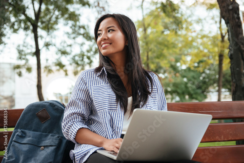 Portrait of young beautiful asian female student with laptop, sitting on wooden bench in park