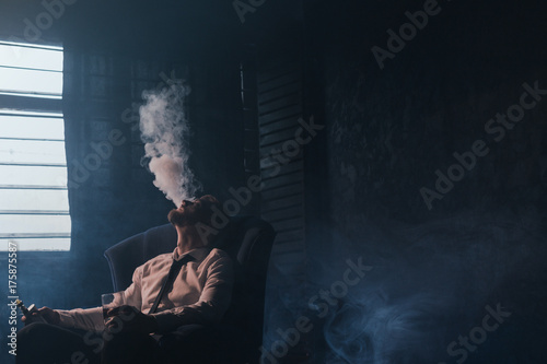 Businessman relaxing with vape and alcohol. Dark room with white atmospheric smoke background, rich people entertainment, private club for luxury life with free space