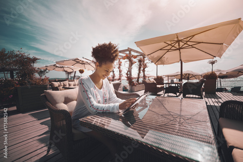 Cheerful curly African American girl is sitting outdoor in a cafe with digital tablet and having online chat with her friend  black smiling lady in street bar nearshore using her digital pad © skyNext