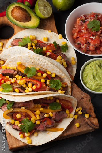 Three Mexican tacos with marbled beef, black Angus and vegetables on wooden Board on a dark stone background. Mexican dish with sauces guacamole and salsa in bowls. Top view