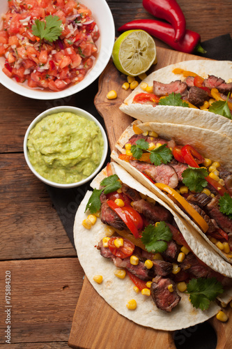 Three Mexican tacos with marbled beef  black Angus and vegetables on old rustic table. Mexican dish with sauces guacamole and salsa in bowls. top view