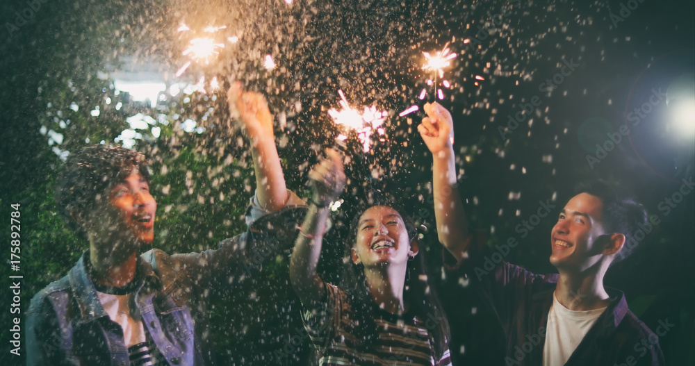 Asian group of friends having outdoor garden barbecue laughing with alcoholic beer drinks and showing group of friends having fun with sparklers on night ,soft focus