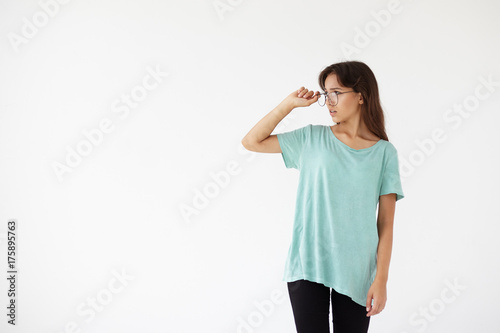 Fashion, style and people concept. Studio shot of stylish teenage girl wearing black jeans and blue oversize t-shirt standing at blank wall and looking sideways, adjusting glasses in trendy frames