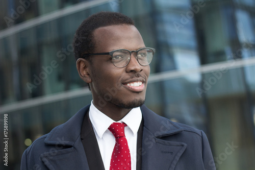 Outdoor headshot of young handsome African businessman pictured in urban environment dressed formally in suit, shirt, tie and coat, looking through eyeglasses to street with optimistic smile