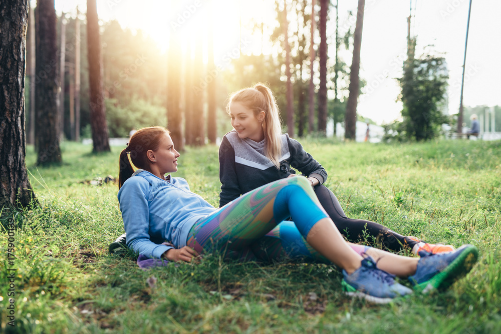 Two sporty girlfriends relaxing, lying on grass and chatting in the forest after jogging together