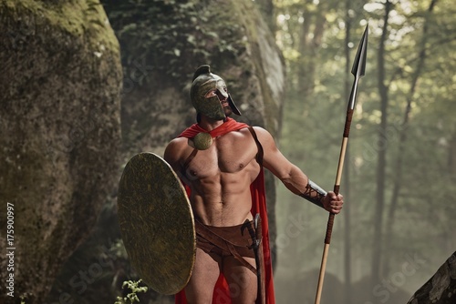 Sportive man with great muscles wearing armor of ancient gladiator and posing with arms on background of rocks and woods.