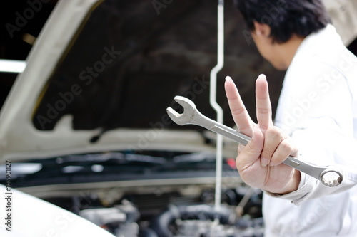 Hands of confident young mechanic holding wrench and raising two fingers up it is shows fight with job against car in open hood at the repair garage.