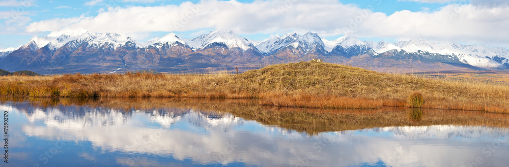 Panoramic view of the mountain ridge of the Eastern Sayan with snow-capped peaks and reflection in the blue water of the river in autumn